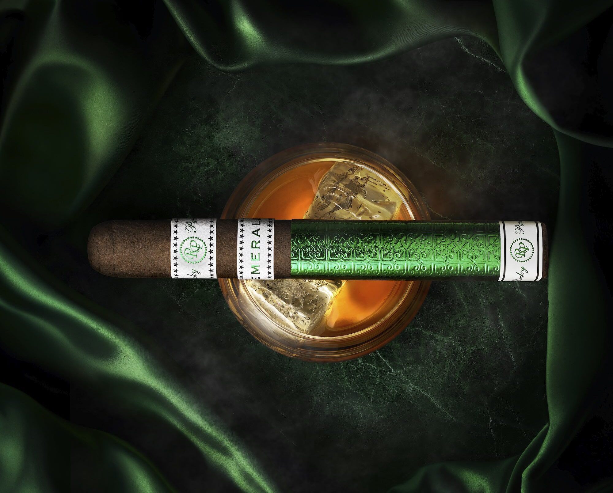 A single rocky Patel Emerald Cigars unlit resting on a rocks glass on a green background
